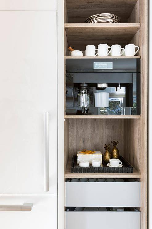 Light gray pull out coffee pod drawers are stacked beneath a shelves holding coffee mugs and a brown shelf holding a Miele coffee machine.