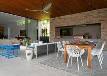 Covered patio features white laser cut outdoor dining chairs at a teak outdoor dining table atop a gray lattice outdoor rug, an outdoor kitchen, a gray wood and rope outdoor sofa with a concrete coffee table, forest armchairs and blue woven stools under a ceiling fan.