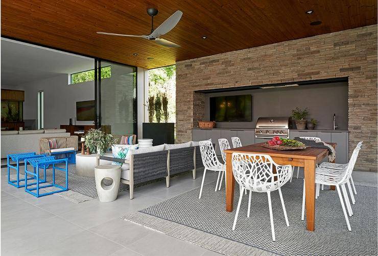 Covered patio features white laser cut outdoor dining chairs at a teak outdoor dining table atop a gray lattice outdoor rug, an outdoor kitchen, a gray wood and rope outdoor sofa with a concrete coffee table, forest armchairs and blue woven stools under a ceiling fan.