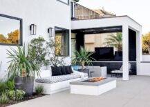 White built-in outdoor sofa upholstered with a gray and white cushion facing a gray granite fire pit in an outdoor patio design. A covered patio features black built-ins with white quartz countertop for a modern appeal.