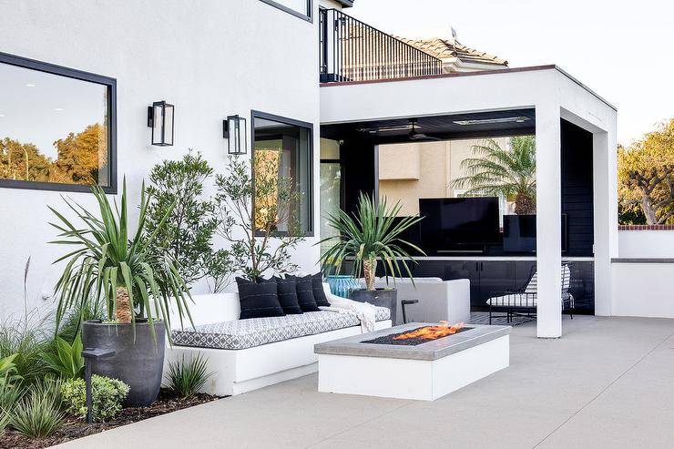 White built-in outdoor sofa upholstered with a gray and white cushion facing a gray granite fire pit in an outdoor patio design. A covered patio features black built-ins with white quartz countertop for a modern appeal.