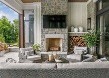Gorgeous covered patio features custom firewood cubbies fixed beside a gray stone fireplace mantel fixed beneath a wall mount TV. Gray wicker lounge chairs sit at on each side of the fireplace.