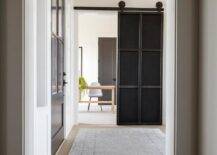 A beautifully styled entryway, a gray rug sits atop a gray wash wide plank wood floor and leads to a home office boasting an industrial metal door on rails.