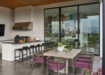 A modern covered patio boasts a polished concrete floor fixed beneath gray and fuchsia dining chairs placed surrounding a gray reeded dining table under a brown plank ceiling. Black low back stools are placed at a white outdoor kitchen island contrasted with a black countertop.
