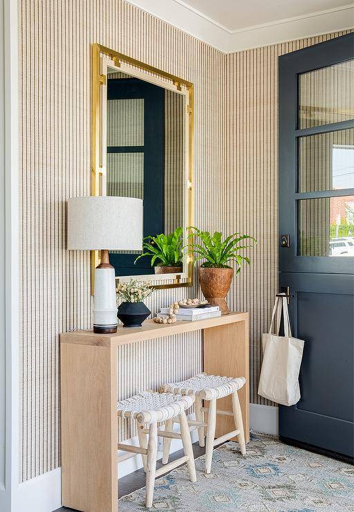 Entryway features white rope benches under a tan oak table, a brass and wood mirror on tan and black vertical stripe wallpaper, a blue and gray rug and a black Dutch door.