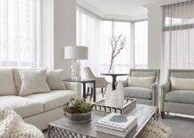 Light themed transitional living room cozies up with an ivory sofa paired with gray accent chairs around a wooden and metal coffee table. A morrocan wedding blanket ottoman under the coffee table boasts texture adding excitement to the simple yet decorated living space.