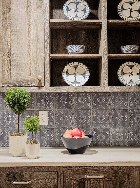 Brass hardware complements rustic kitchen cabinets fixed against white and gray mosaic tiles beneath reclaimed wood shelves.