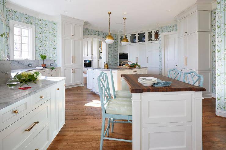 White and blue kitchen features blue and green wallpaper, a walnut top center island with sky blue bamboo counter stools decorated with green Greek key cushions, white cabinets with gray marble countertop and brass pulls and brass and white pendants over a second island.
