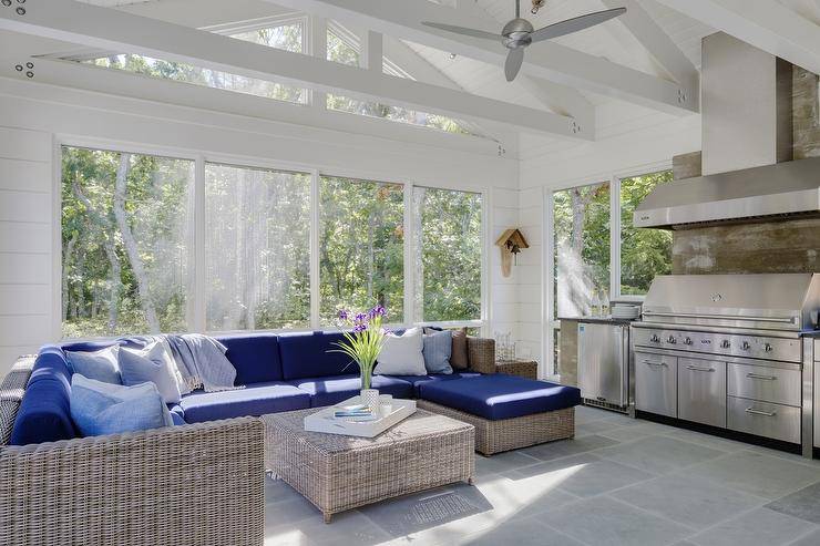 Beautiful sunroom with a white truss ceiling accented with a silver ceiling fan features a wicker sectional fitted with blue cushioned seats around a wicker ottoman as a coffee table atop gray pavers leading to the sunroom kitchen with a vent hood over plank backsplash and barbecue next to a stainless steel mini fridge.