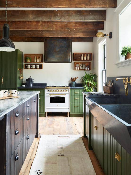 Cottage style kitchen features a white and gold French stove with a black metal hood flanked by wooden shelves over green paneled cabinets, a honed black marble dual apron sink with antique brass faucets and a black distressed paneled kitchen island with marble countertop lit by glossy black pendants with wooden ceiling beams.