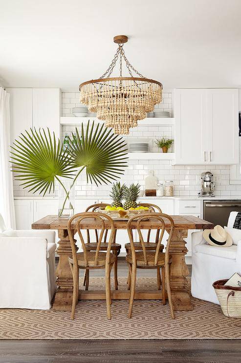 Beige beaded chandelier over a small reclaimed wood dining table finished with two white slipcovered dining chairs and French bistro chairs on a jute hexagon rug. This open concept kitchen and dining space uses the area practically and decoratively balancing warm and cool tones.