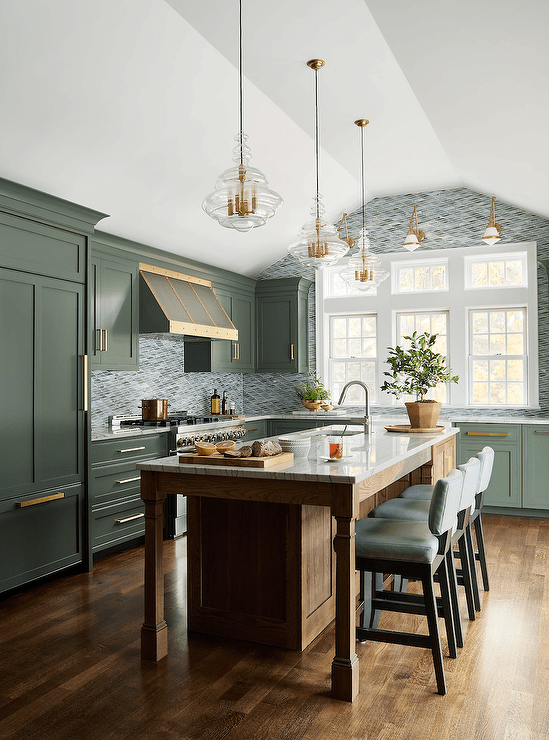 Green kitchen with a brown oak island topped with gray striped marble lit by glass and brass pendants features green leather stools, green cabinets with brass pulls and dark wooden floors.