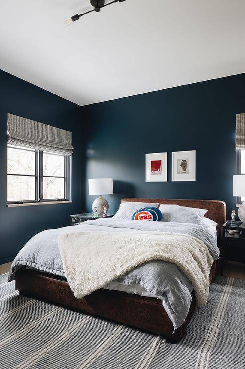 Bold blue walls accent a charming boy's bedroom featuring an ivory and gray rug placed beneath a brown leather bed dressed in light gray bedding and complemented with a white plush throw blanket and a cubs pillow. The bed is placed beneath side-by-side framed art and between glass and metal nightstands.