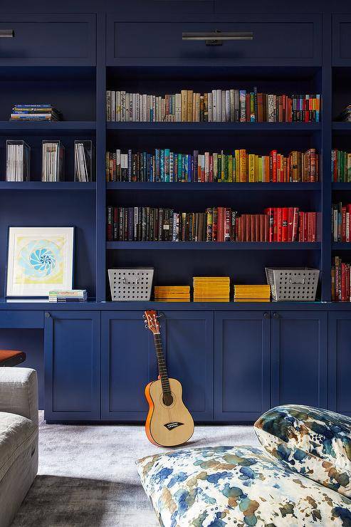 Home office features bold blue bookshelves with books organized by color lit by bronze picture lights and paint splatter accent pillows.