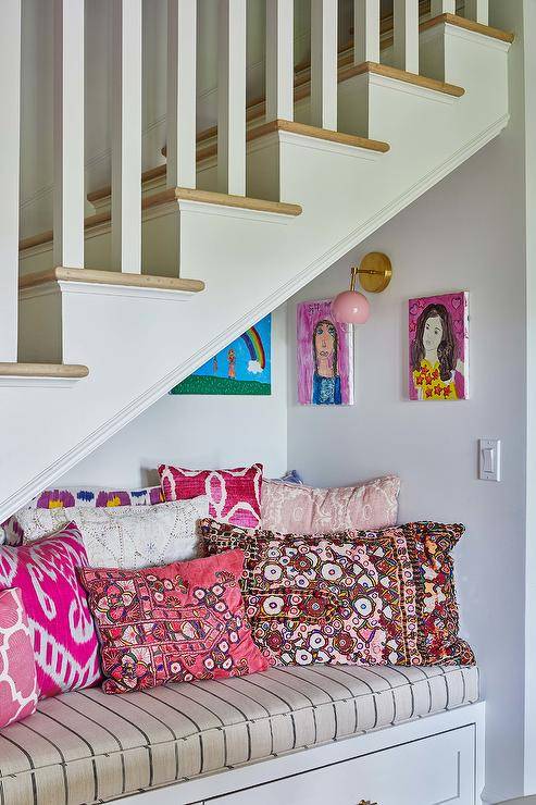 Beneath a staircase, a cozy reading nook boasts a white built-in bench accented with a French burlap cushion and layered pink pillows positioned beneath hung children's art lit by a pink sconce.