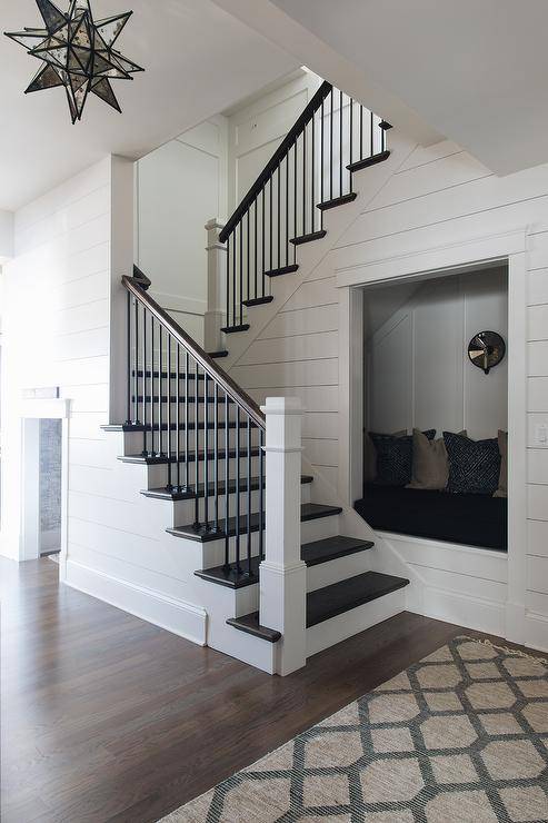 Recessed into a wall, a reading nook is fitted with a built-in seat topped with a black cushion located against a white board and batten trim. The nook is framed by white shiplap and is fixed facing a white staircase contrasted with dark stained wood treads matched with a dark stained wood railing.