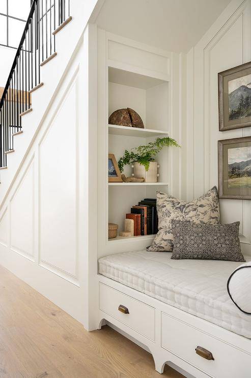 Cozy reading nook with built in shelves over a bench with a gray cushion and drawers with vintage brass pulls sits under a staircase.