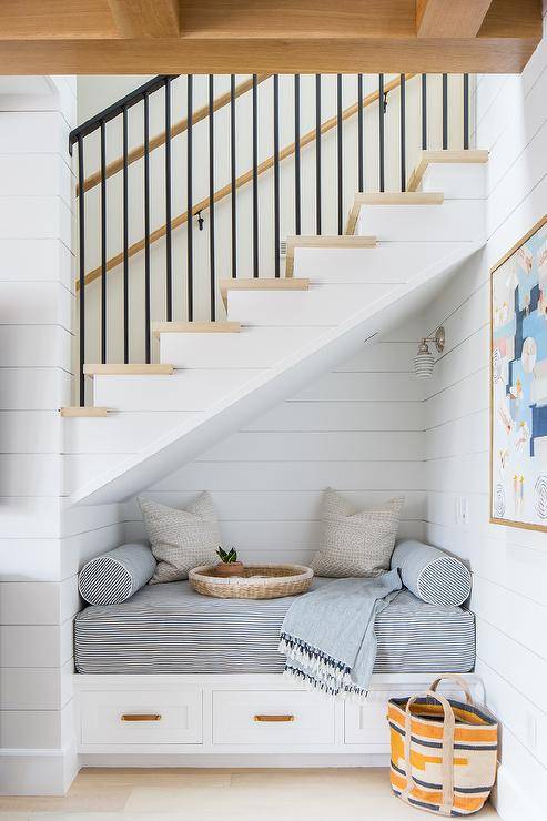 Beneath a staircase boasting blond wood treads, a white shiplap reading nook is fitted with a white built-in bench. The bench boasts drawers and a blue pinstripe cushion.