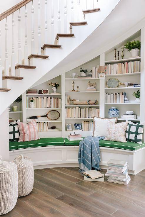 A curved built-in bench topped with kelly green cushions is fixed beneath a winding staircase and under white built-in bookshelves. The staircase boasts stained wood treads and a stained wood handrail fixed against white spindles.
