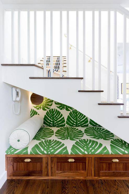 Built-in reading nook bench under a white staircase boasting a lovely and vibrant white and green palm leaf print cushion. A white lumbar pillow joins the cozy setting atop wood built-in bottom drawers with vintage brass cup pulls.