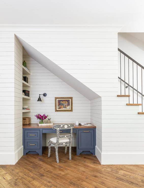 Beneath a staircase, a blue desk with a butcher block top sits on a diagonal wood floor and is built-in beneath an art piece hung from a wall covered in shiplap trim. A black and white bone inlay chair sits at the desk, while a white shelves are built-in to the side of the desk and lit by an oil rubbed bronze sconce.