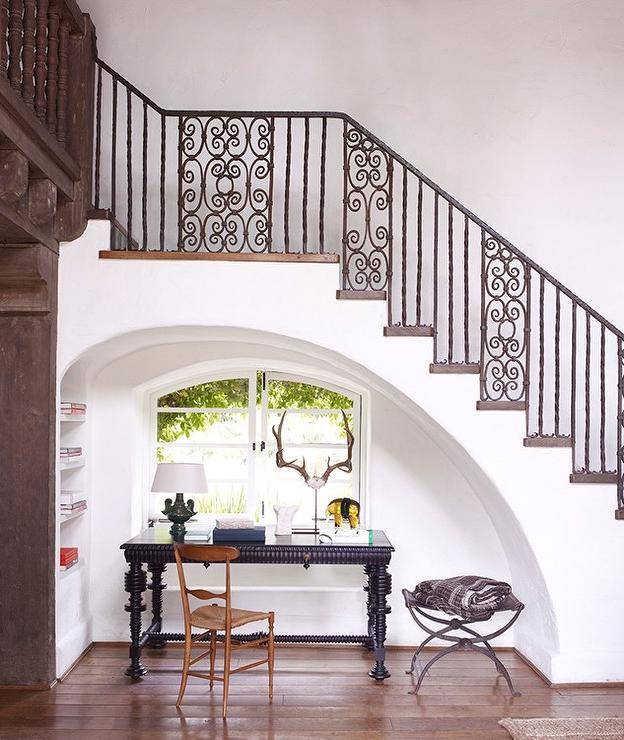 Mediterranean home features an arched alcove, placed under a wrought iron staircase, filled with a black desk and a wooden chair placed under arched casement windows situated next to built in shelving.