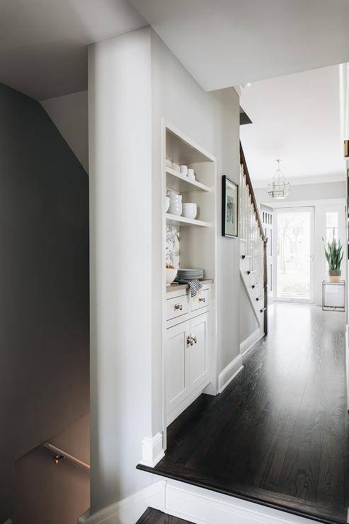 Built-in to the side of a staircase, a butler's pantry nook features white cabinets adorned with vintage hardware and fixed beneath stacked white shelves fixed against white and gray wallpaper.